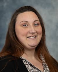 Brittany Yenior, Administrative Secretary, profile portrait. She has long brwon hair, wearing a black cardigan over a white and black floral top and a grey background for the picture