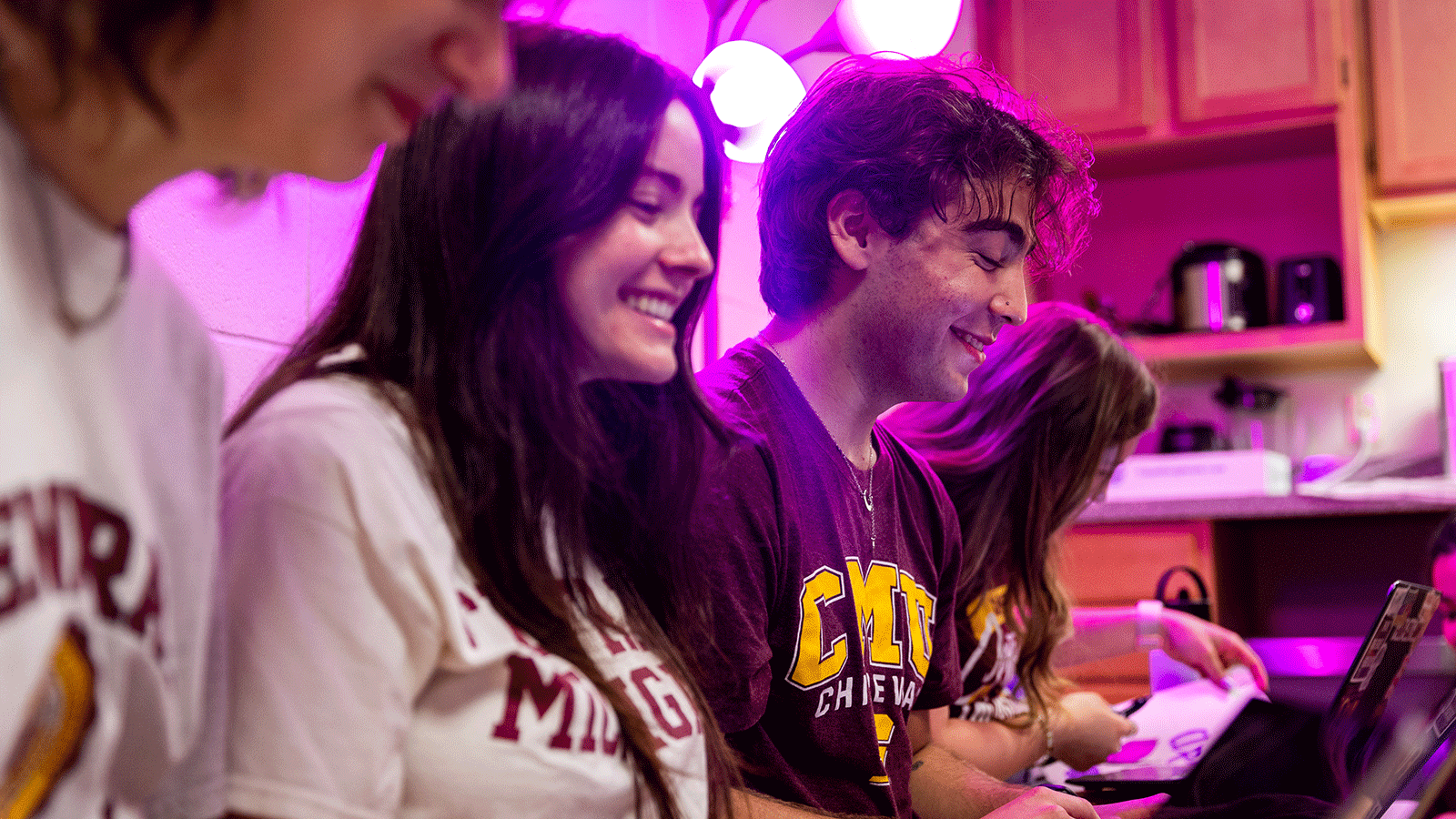gender inclusive housing Image of five students sitting on a couch in a residence hall room as they wear CMU t shirts and are working on their laptops, smiling. There is a pink light behind them.