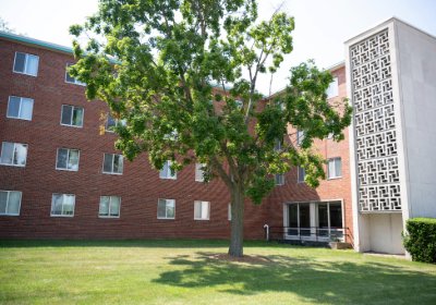 Sweeney Hall's red brick exterior with tall windows, shaded by a large tree and bordered by a lawn.