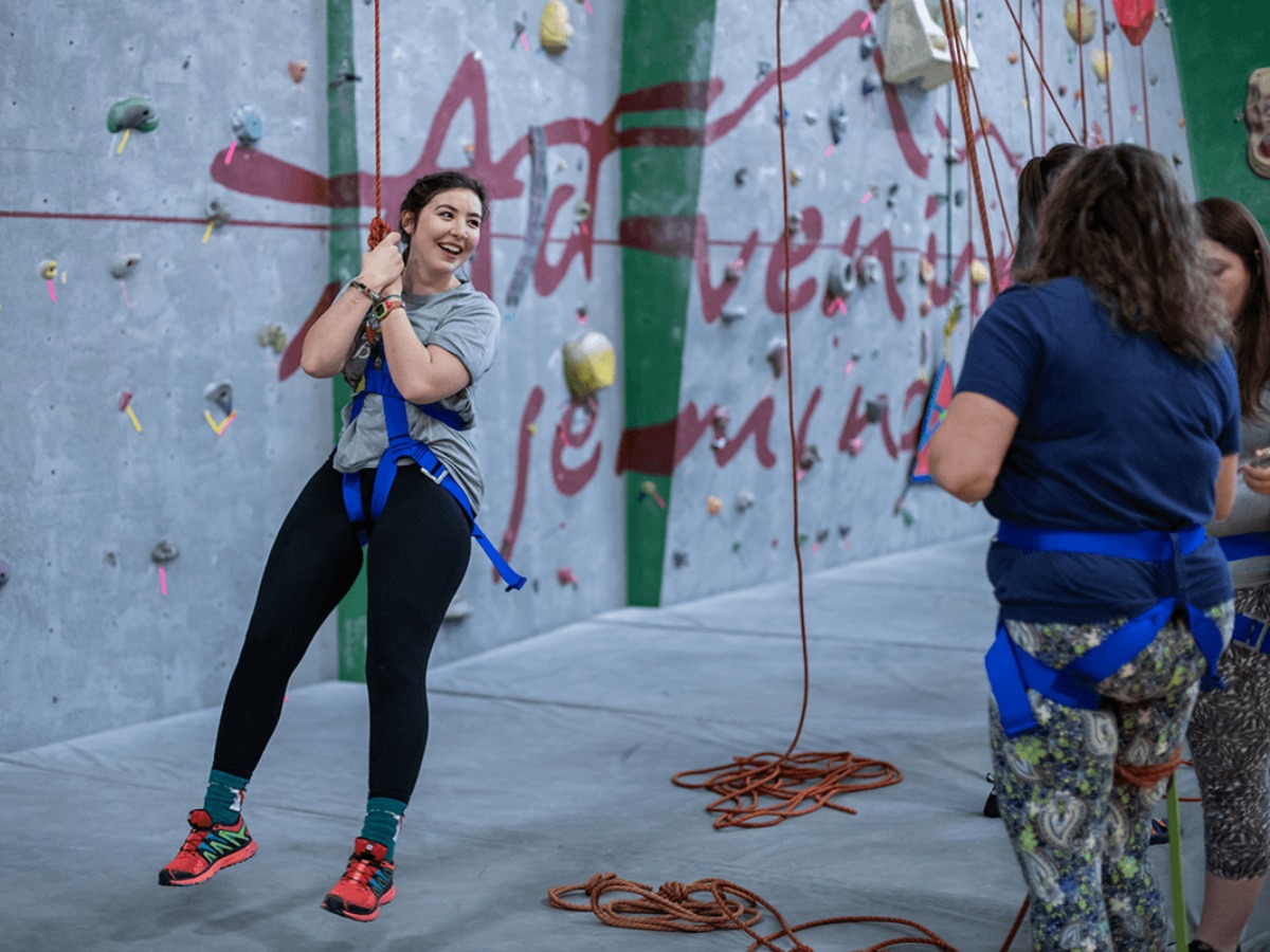 student in a blue harness smiles at the bottom of a rock wall