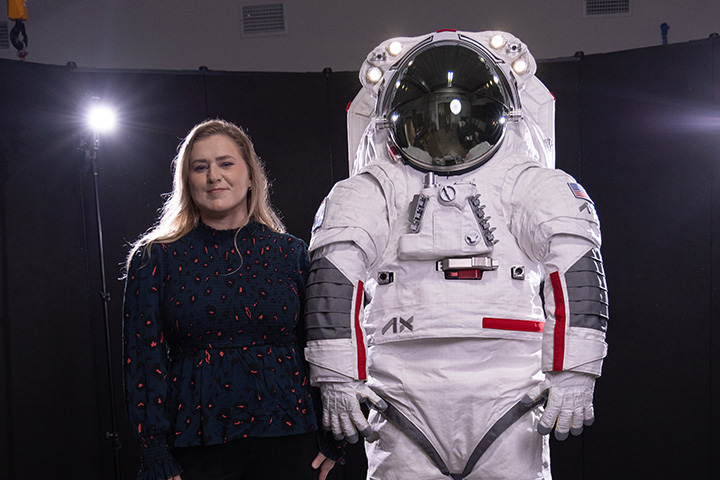 Falanne Jenkins poses in a dark suit next to a spacesuit. Photography lights and a black curtain backdrop are behind them.