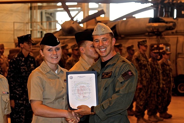 A woman in a Navy uniform receives a certificate from a man in a Navy aviator's uniform. They are standing in front of a helicopter and a large group of other servicemembers. The servicemembers are standing at attention.