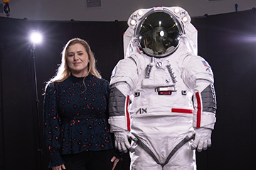 A woman in a dark shirt standing next to a model of a spacesuit. There are photography lights behind them and a dark curtain backdrop.