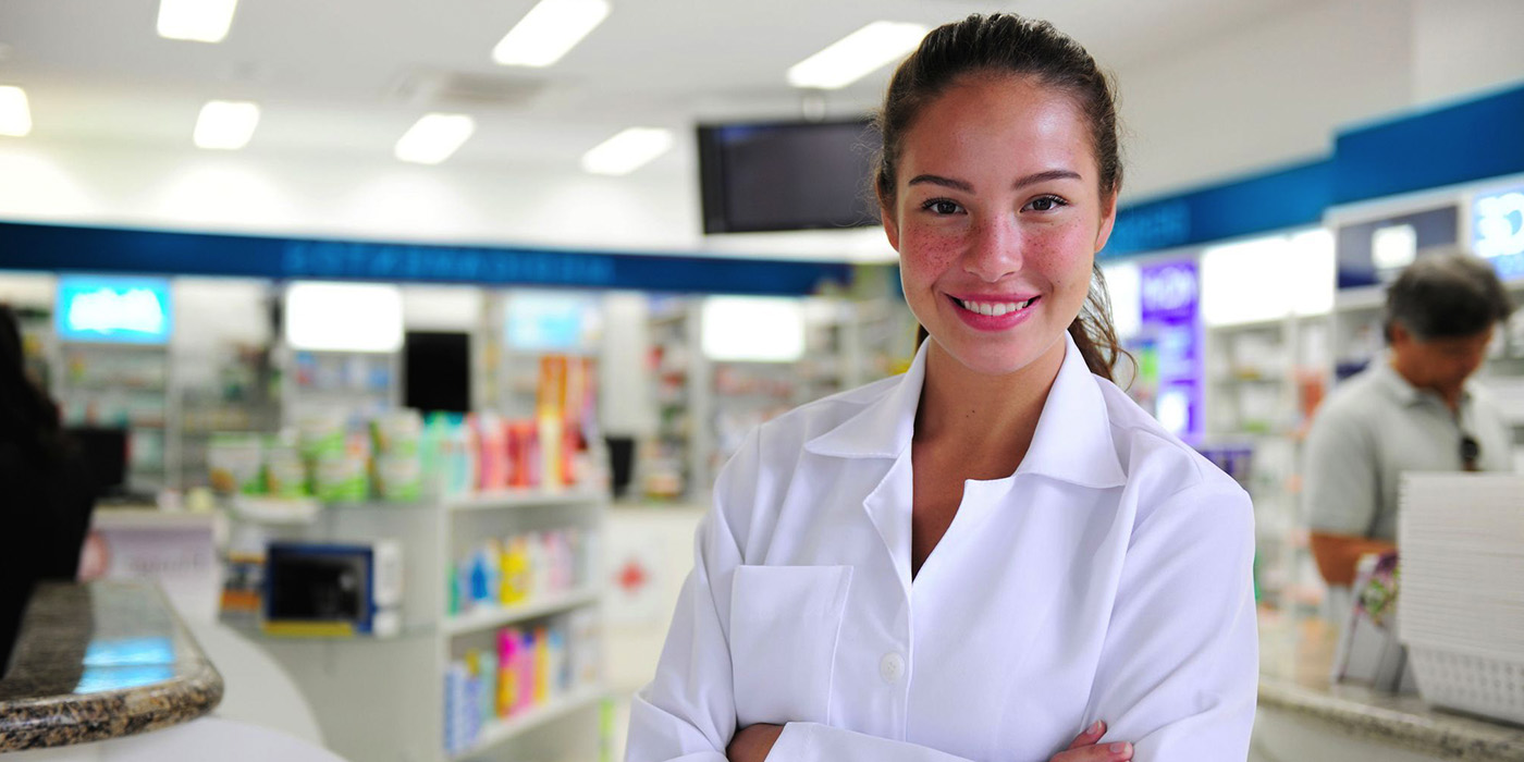 A pharmacy technician with dark hair wears a white lab coat.