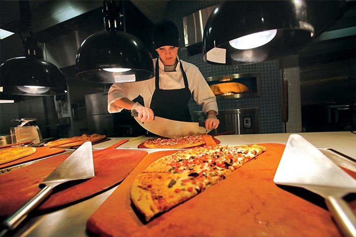 A person in a chef's hat slices a pizza with a specialty cutter. Behind them is a pizza oven. In the foreground is a half of a pizza on a serving tray.