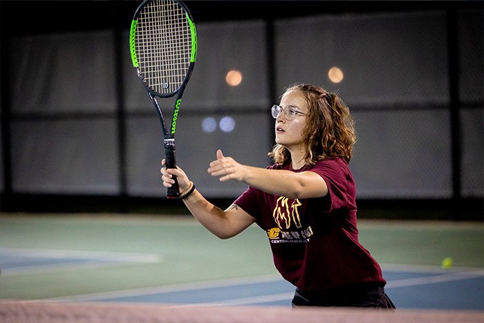 A student, looking focused, holds a tennis racquet up to return a ball across a net.
