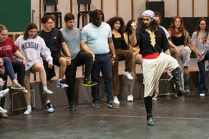 A group of students stand in a line with one foot raised mid-air, learning the steps of Dabke from an instructor