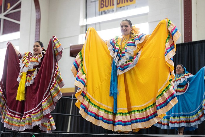 Three dancers perform folklórico on stage with their arms outstretched in colorful dress.