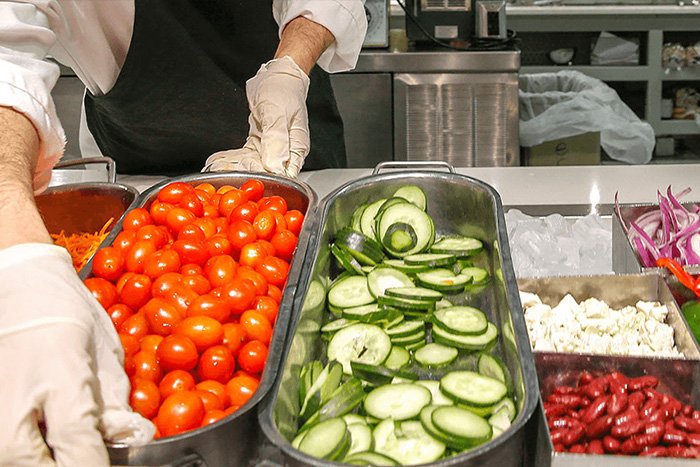 A spread of fresh food including tomatoes, sliced cucumbers and beans. A pair of gloved hands adjust the tray of tomatoes.