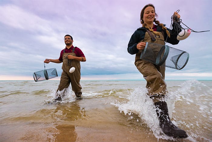 Two students wearing waiters and holding fishing nets walk through the surf on Beaver Island.