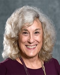 Pam Murray is an Advancement Board member and she is wearing a maroon top and a maroon and gold necklace and she has shoulder-length curly hair.