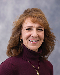 Jean Brown is an Alumni Board member who holds Emeritus status and she is wearing a dark red coat with a maroon turtleneck with shoulder-length autumn brown hair.