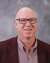 Alumni Board member Jonathan Eadie standing in front of a gray background and he is wearing a maroon suit jacket and a maroon plaid shirt.