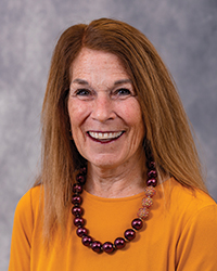 Linda Leahy is an Alumni Board member who is wearing a gold top and a beaded necklace and she has long autumn hair.