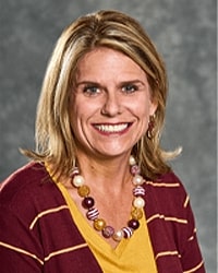 Alumni Board member Norma Eppinger wearing a maroon and gold striped cardigan and yellow shirt with a large beaded necklace and she has shoulder-length blonde hair.