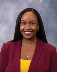 A close-up of a woman smiling at the camera. She has black hair and wears a maroon blazer over a gold tank top. She wears gold earrings and a gold necklace.