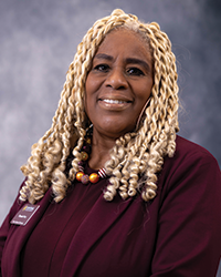 A woman with blonde hair smiling at the camera for a headshot photo in front of a grey background. She is posed at an angle and wearing a maroon shirt, blazer, necklace, and hoop earrings.