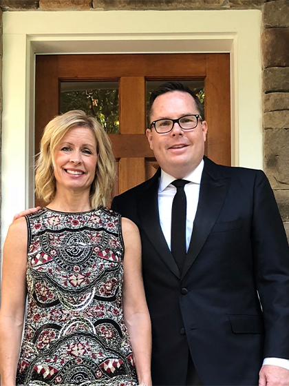 Bob and Lisa Oros 420x560 A man with glasses in a black suit, white dress shirt and black tie stands next to a woman in a black and red paisley dress. In the background is a door to a building.