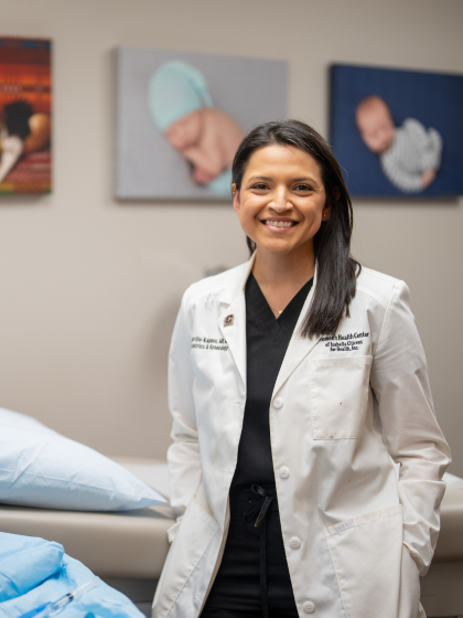A woman with dark hair in a white doctors lab coat with two portrait pictures of babies on the wall behind her.