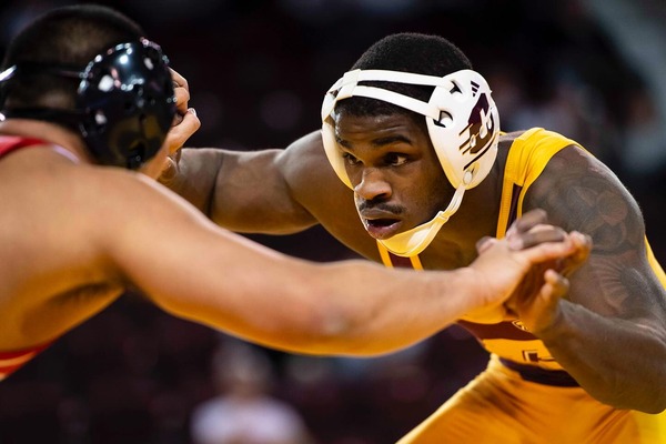 A Central Michigan University wrestler in a gold singlet and protective headgear focuses intently while engaging an opponent.