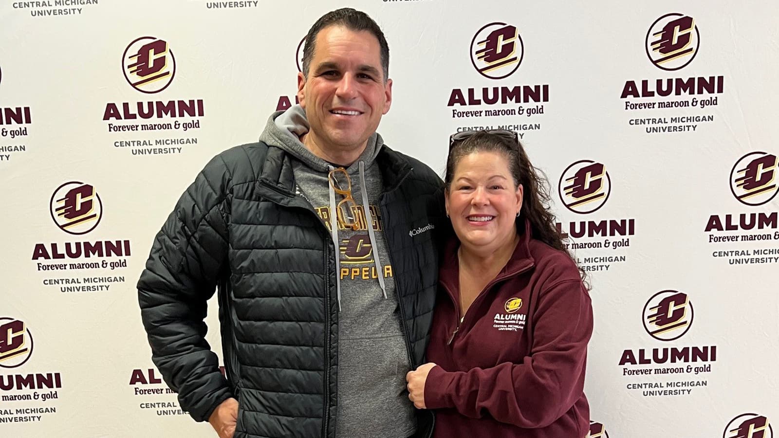 A man and woman wearing Central Michigan University tops stand together in front of a CMU Alumni step and repeat background wall.