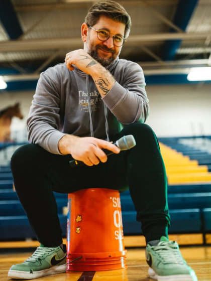 A man with short dark hair and glasses sits on an orange bucket inside a gym while holding a microphone.