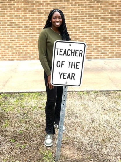 Akayla Joseph, '18, standing behind a teacher of the year sign in a green shirt and black pants.