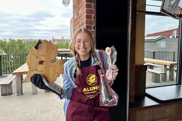 A woman wearing a maroon CMU alumni apron holds a wooden cutting board shaped like Michigan and a wooden spoon with an Action C.