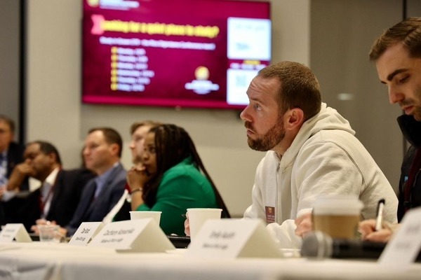 Several individuals are attentively listening to a presentation while seated at a long table with name cards.