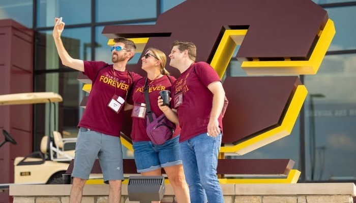 Three Central Michigan University alumni in maroon shirts taking a selfie in front of large Action C signage.
