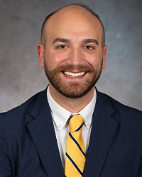 Headshot of James Nuse for the 2024 10 Within 10 Alumni Awards and he is wearing a dark navy suit with a blue and yellow striped tie and he is smiling with teeth and has a brown beard.