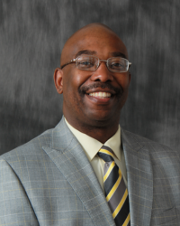 A man with a bald head wears eyeglasses, a grey suit and striped tie.