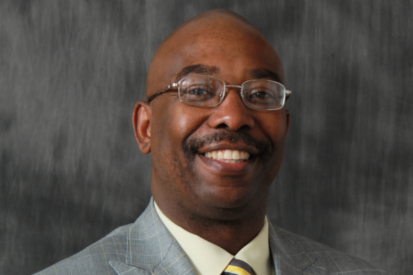 A man with a bald head wears eyeglasses, a grey suit and striped tie.
