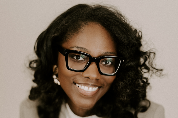 A woman with dark, curly hair wears glasses and a tan blazer over a white shirt.