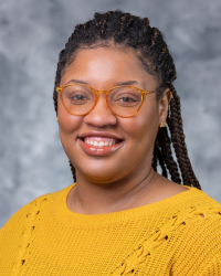 Jasmine Hall 200x250 A woman with black braided hair is wearing glasses and a yellow sweater.