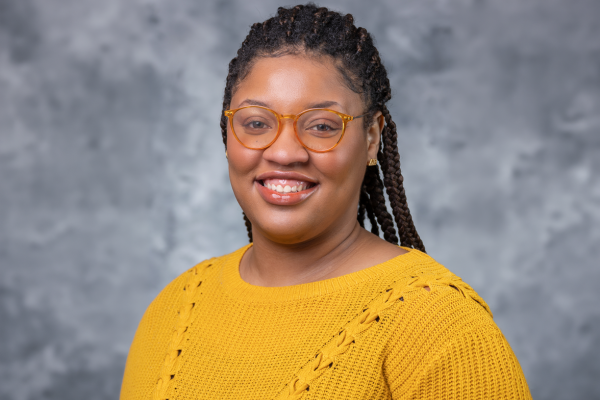 A woman with black braided hair is wearing glasses and a yellow sweater.