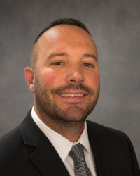 Headshot of a man with a short beard and short dark hair is wearing a black suit, dark tie and white dress shirt.