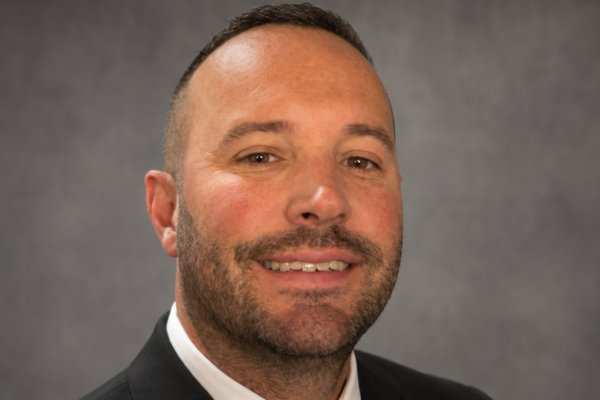 Headshot of a man with a short beard and short dark hair is wearing a black suit, dark tie and white dress shirt.