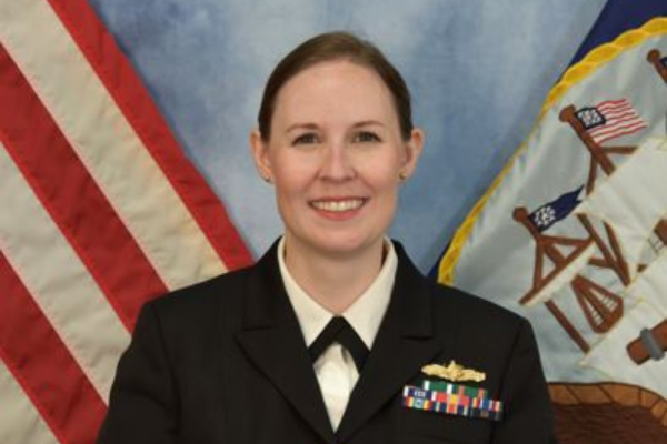 A woman in a military uniform smiles with one hand on top of the other in front of her. A US flag is over her left shoulder and a white flag with various brown symbols including a small US flag on it is over her right shoulder.