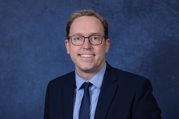A man with glasses and short brown hair smiles while wearing a dark suit, navy blue tie and light blue shirt.