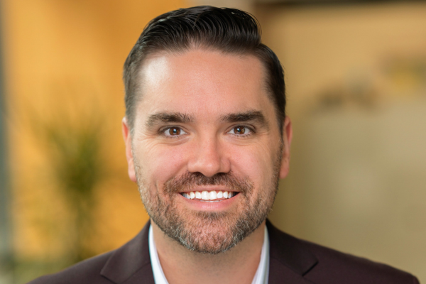 Headshot of a man with short facial hair and dark hair wearing a dark suit and white dress shirt.