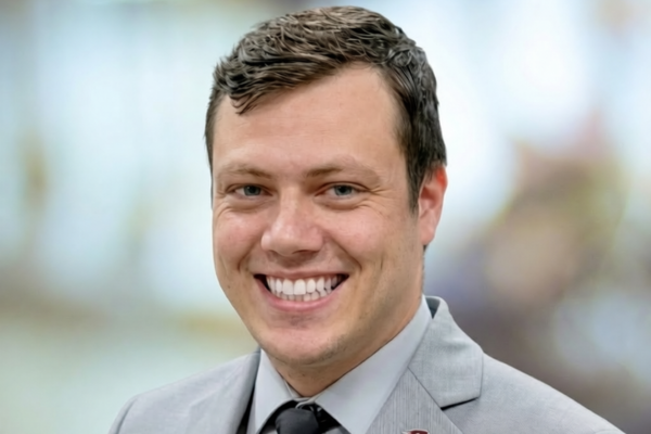 A man with glasses and short brown hair smiles while wearing a dark suit, navy blue tie and light blue shirt.