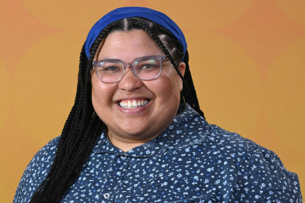 A woman wearing glasses with long black hair, a royal blue headband and a blue shirt with light blue spots all over it is smiling at the camera.
