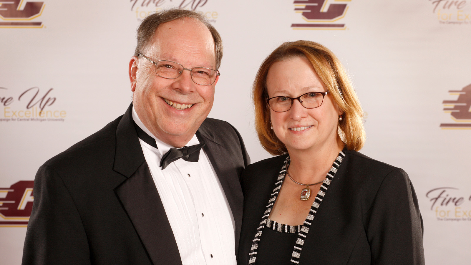 Oppermans portrait 1600x900 A man in a tuxedo smiles and poses next to a smiling woman wearing a black dress.
