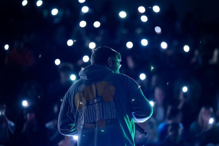 Back view of Joe Vercellino on a dark stage wearing a hoodie during a Lion Heart Experience event, facing an audience lit by phone flashlights.