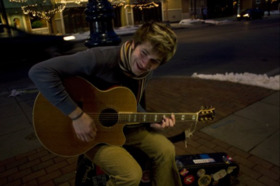 Young Joe Vercellino playing an acoustic guitar while sitting outdoors on a Mount Pleasant sidewalk at night with snow on the ground.