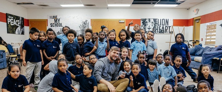 Joe Vercellino smiling while surrounded by a large group of young elementary students sitting and standing together in a school classroom.