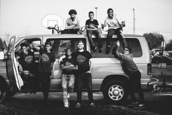 Black and white photo of Joe Vercellino standing with a group of young men, posing on and around a passenger van while wearing matching Lion Heart graphic t-shirts.