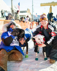 Keaton and Erika Loebrich smiling and kneeling in the snow with two dogs wearing protective booties during a dogsledding race event.
