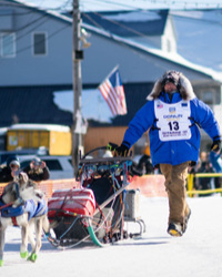 Dogsled racer Keaton Loeblich '22, wearing a bright blue parka with a fur hood and the number 13 bib, runs alongside his sled and dog team on a snowy track.